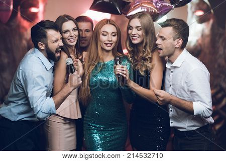 Young people in the club dance and sing. A man in a white shirt is holding a microphone and singing. His friends are singing along with him. They are in a trendy modern nightclub.