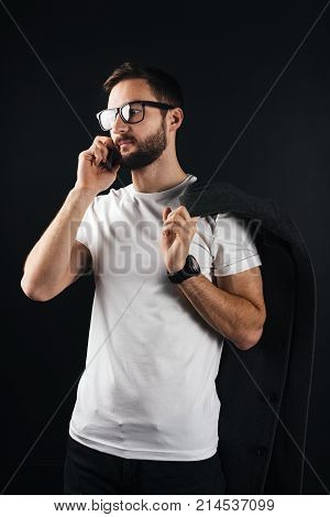 Young Bearded Hipster In Glasses Talking On Smartphone Standing In White Blank Thirt And Keep Coat O