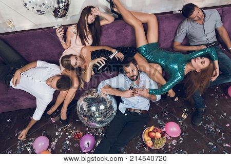 Young people are resting after a tumultuous party in a nightclub. They lie chaotically and effortlessly on the lilac sofa. Around them are confetti and colorful balloons.