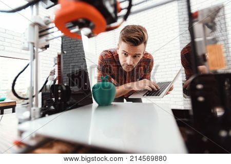 A man with a laptop in his hands controls the process of printing a 3d printer. 3d printer has printed model of an apple. The man controls the process.