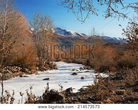 Sunny autumn mountain valley with a river stream and shores with yellow dry grass on the background of snow peaks and ice glacier cloudy sky. Ice almost got up.