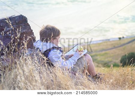 Boy reading book outside on the top of the hill, snacking with healthy apple, unschooling education