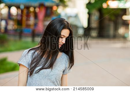 Young thoughtfull female in gray shirt with long brunette hair looking down in the street. Close up portrait