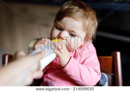 Cute adorable baby girl drinking formula milk. First food for babies from bottle holding by mohter. New born child, sitting in chair of domestic kitchen. Healthy babies and bottle-feeding concept.