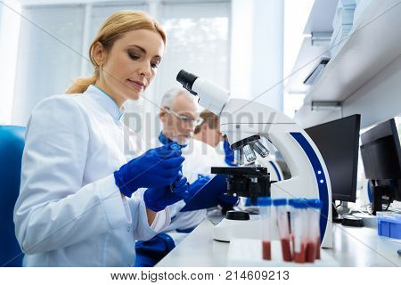 Working with blood. Concentrated blond young researcher sitting at a microscope and holding test tubes while wearing a uniform in the lab and two other researchers working in the background