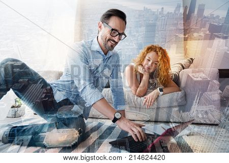 Warm atmosphere. Positive clever responsible man looking happy while sitting on the floor and showing the results of his work to a cheerful smiling woman