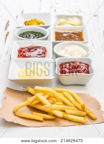 French fries on a piece of crafted paper with different sauces in white sausers on the white wooden background with cracking paint