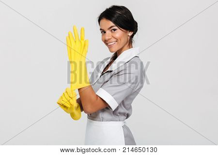 Portrait of beautiful brunette housekeeper in uniform wearing yellow rubber gloves while smiling and looking at camera, isolated on white backgrund