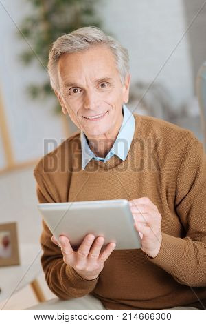 Getting updated. Cheerful senior man looking into the camera with his eyes full of positivity and smiling while sitting in a chair and working on his touchpad at home.