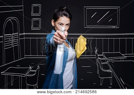 Gun down with purity. Pleasanrt beautiful woman holding cleaning spray and doing spring cleaning while lookign at you