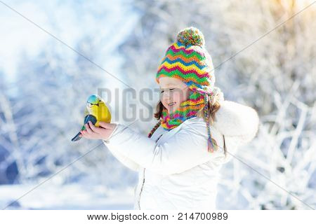 Child feeding tit bird in winter park. Kids feed birds in snowy forest. Little girl with titmouse. Kids watch wild animals. Family Christmas vacation. Children play in snow. Outdoor and wildlife fun.