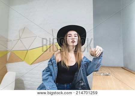 Portrait of a stylish woman sitting in a cafe posing on the camera. The hipster girl poses in the cafe with a cup of coffee in his hands
