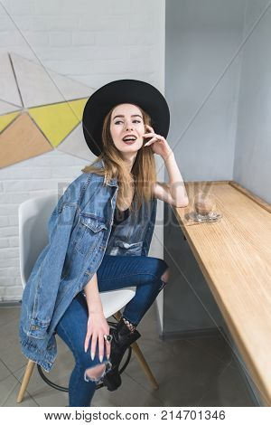 A stylish woman poses to the camera in a cozy cafe for a cup of hot drink near the window. Portrait of a hipsters girl in a cafe