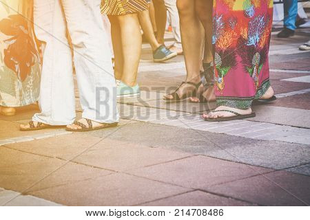 Group of casual men and women standing on paving in a low angle view of their feet in sandals jeans shorts and caftans or long summer skirts with the sun glow in the left corner