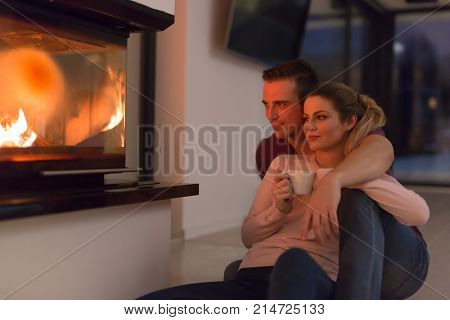 Young romantic couple sitting on the floor in front of fireplace,talking and drinking tea on cold winter night at home