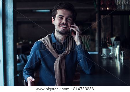 Ok I am waiting for you! Handsome young man talking on mobile phone and looking away
while sitting at bar counter