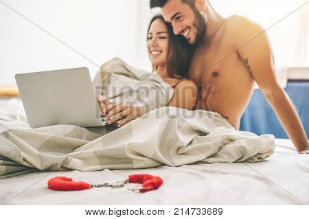 Happy young couple lying on a bed with computer - Beautiful married couple watching role games sex video on laptop laughing together - People sexual technology concept - Soft focus on sheets