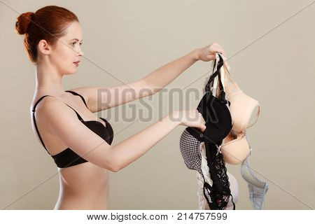 Bosom concept. Slim attractive red hair woman wearing black underwear holding many bras in hand choosing witch bra to wear