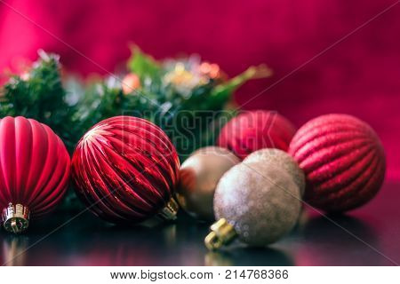 Red and gold Christmas ball ornaments are placed on a black reflective table top surface. The ball ornaments are different sizes and are shiny and matte colored. The ornaments reflect off the table. Behind the ornaments is a green Christmas wreath and a r