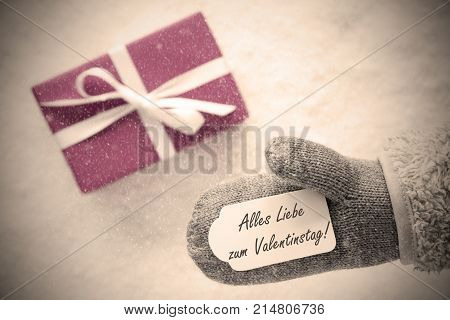 Glove With Label With German Text Alles Liebe Zum Valentinstag Means Happy Valentines Day. Pink Or Rose Gift Or Present On Snow In Background. Greeting Card With Snowflakes And Instagram Filter