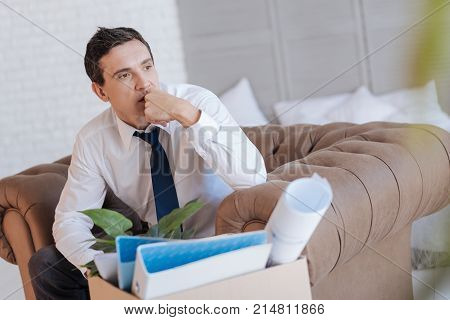 Difficult decision. Clever experienced specialist sitting in an armchair with a box of personal items by his side and looking into the distance while thinking of leaving his work
