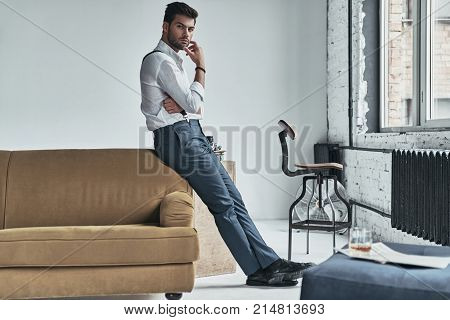 He is looking into your heart. Full length of thoughtful young man in white shirt and suspenders looking at camera and keeping hand on chin while leaning on the sofa