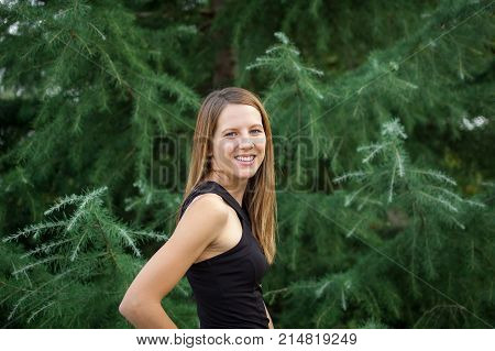 Pretty Beautiful Smiling Woman in Black Shirt with Long Shine Hair Posing near Spruce Tree During Summer Vacation