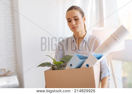 Losing job. Sad quiet young woman looking calm and frustrated while coming home with a heavy box full of personal items after losing her job