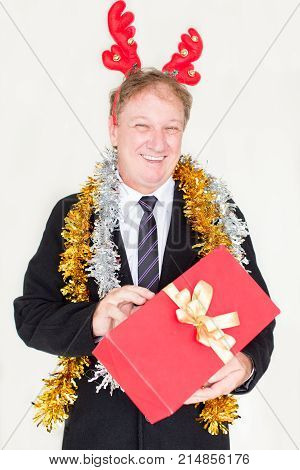 Content funny man in reindeer headband holding Christmas gift and looking at camera. Cheerful pleased handsome businessman in coat ready for gift-giving in office. Christmas joy concept