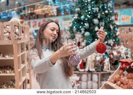 Cheerful happy customer girl choosing gifts for Christmas and New Year. She is taking red Christmas tree decoration balls. difficult decision. Various choice