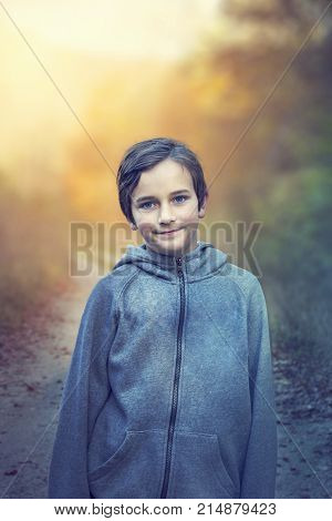 Young boy with blue eyes walking in forest on sunset. Portrait of kid (boy) in forest road (path) with sunlight in autumn time. Boy smile and posing to camera in park on sunset.
