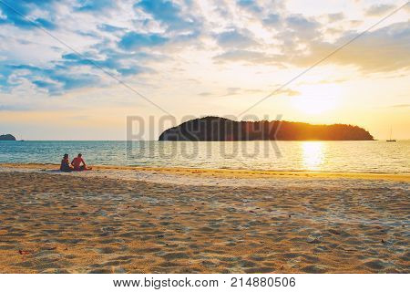 Pantai Tengah Beach at colorful sunset, Langkawi Island, Malaysia. Beach sunset is a golden sunset sky with a wave rolling to shore as the sun sets over the horizon. People relaxing on paradise beach