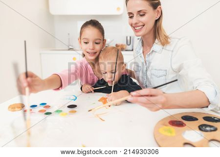 Mother and children are engaged in drawing. They have fun in the kitchen. The girl is holding her younger brother in her arms. A mother of two children teaches drawing.