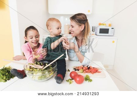 Mom and daughter have fun while preparing a salad. They are in a bright kitchen. Baby boy helps mom and sister to prepare salad.