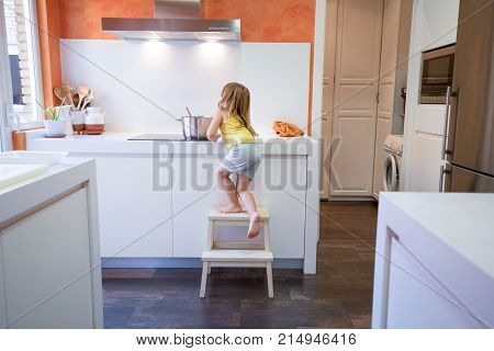 Four years old blonde child climbing on stool o ladder to cook in electrical cooktop with a saucepan alone in the kitchen