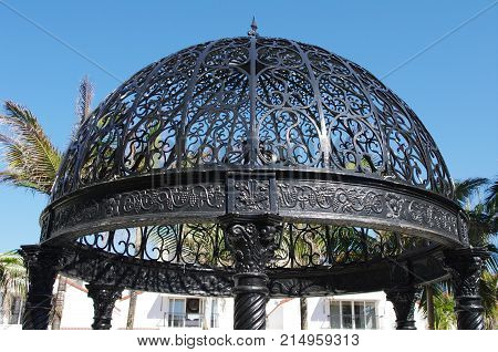 Black steel gazebo dome silhouetted against blue sky with intricate engraving around the edge