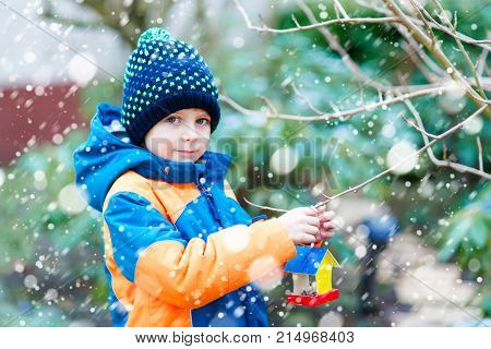 Little kid boy feeding birds in winter. Cute happy preschool child hanging colorful selfmade bird house on tree on frosty cold day. Preschooler in colorful wam clothes. Nature, empathy with animals.