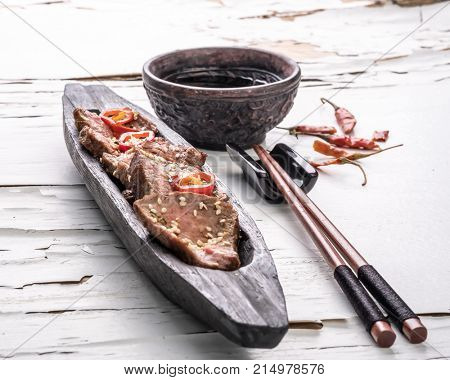 Slices of oriental spicy meat with sesame seads and pepper on long dark wooden plate with chopsticks and bowl of sauce on the white surface with cracked paint. Close up.