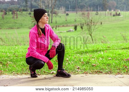 Young woman analyzing the track before running on a cold winter day on the training track of an urban park. Female athlete wearing pink windbreaker, beanie, gloves and running tights