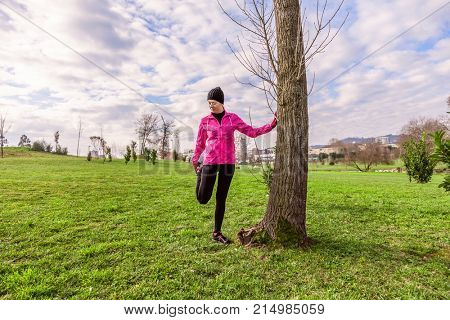 Young woman warming up and stretching the legs before running on a cold winter, autumn of fall day in an urban park. Female athlete wearing pink windbreaker, beanie, gloves and running tights