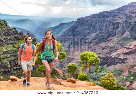 Young happy hikers people walking on Hawaii Waimea Canyon Trail, Kauai island, USA. Asian woman and man couple trekkng in scenic mountain background. Hiking adventure in nature.