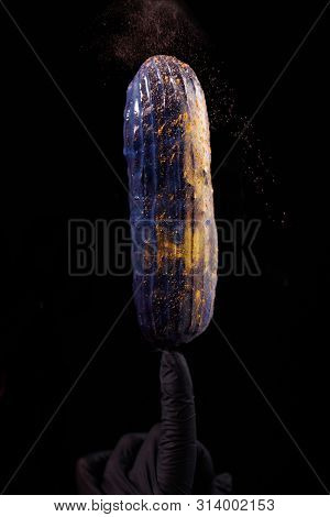 Beautiful Bright Eclair With Blets On A Dark Background. Hands Pastry Chef In Black Gloves.
