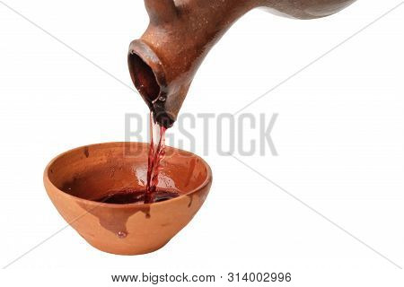 A Red Wine Is Pouring From Traditional Georgian Wine Jug In To The Clay Cup, Isolated White Backgrou