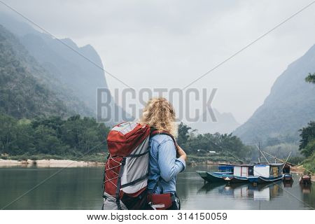 Caucasian Blonde Woman With Backpack Stands On Boat Pier In Muang Ngoi Village, Laos