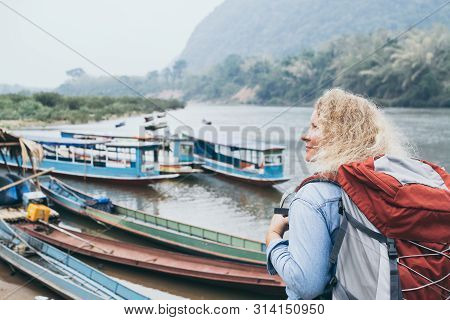 Caucasian Blonde Woman With Backpack Stands On Boat Pier In Muang Ngoi Village, Laos