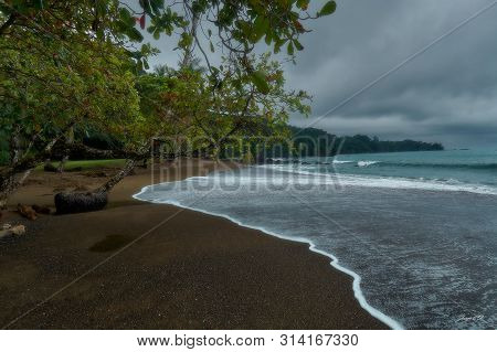 Paradisaical Beach With Some Waves Partly Covering The Dark Sand, Some Tropical Trees & A Stormy Wea