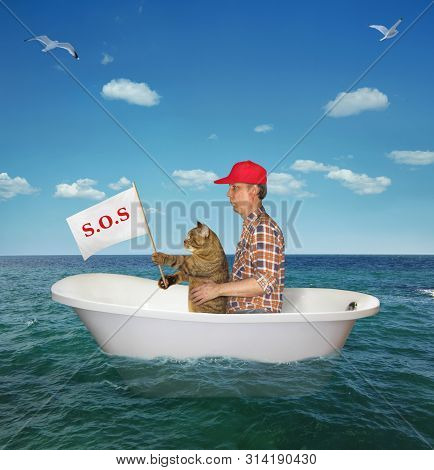 The Man In A Red Cap With His Cat Are Drifting In A Bathtub On The Sea After A Shipwreck. The Cat Ho