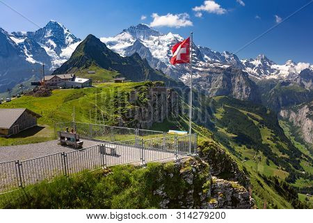 Swiss Flag Waving And Tourists Admire The Peaks Of Monch And Jungfrau Mountains On A Mannlichen View