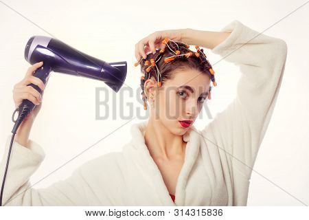 Young Woman In Bathrobe Drying Her Hair With Curlers On Head, Isolated On White Background