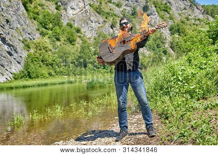 The Musician Plays A Burning Acoustic Guitar Against The Backdrop Of A Mountain River On A Sunny Sum